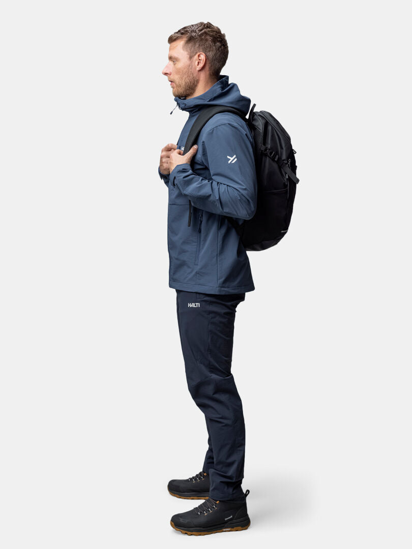 Mann in Halti Finholma X-Stretch Jacke, blauer Hose und schwarzem Rucksack vor schlichtem Hintergrund.