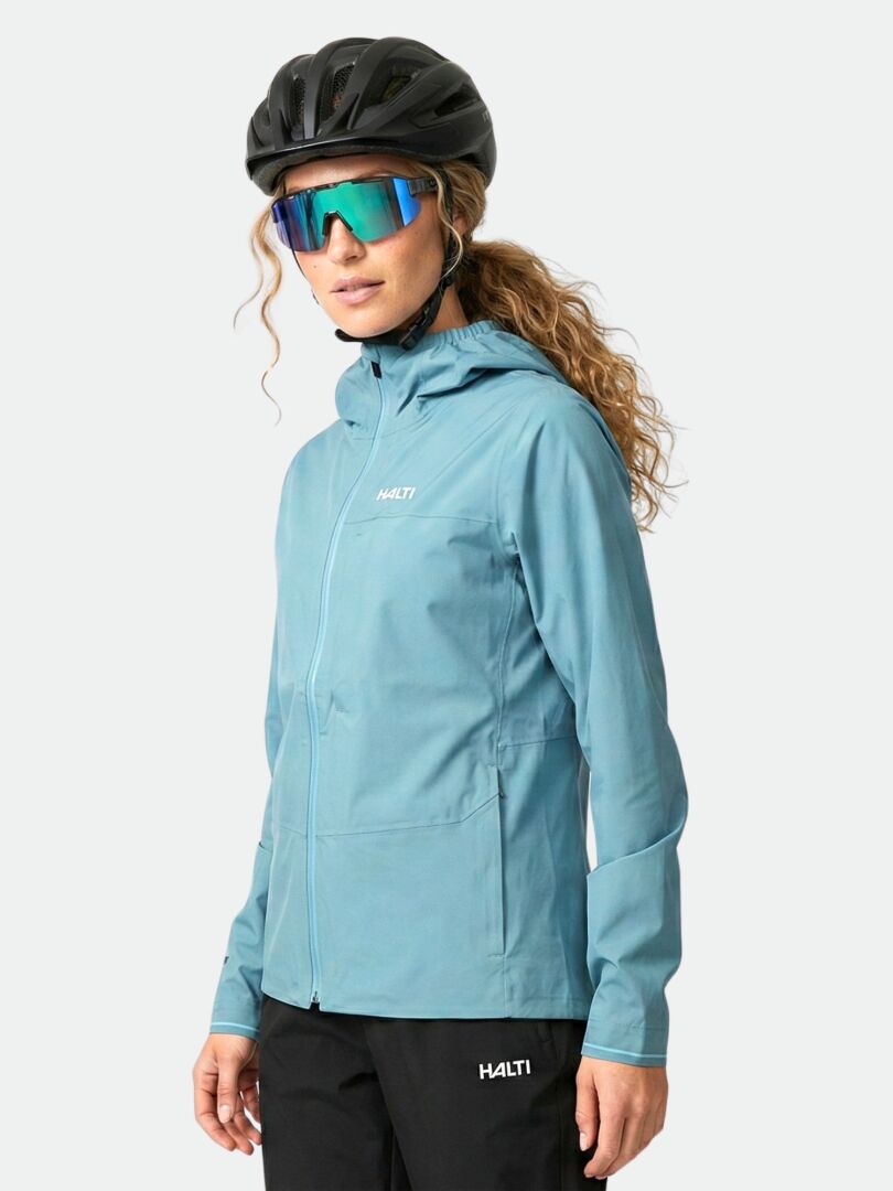 Person, die eine blaue Halti Taival DX 3L Jacke Damen, einen schwarzen Helm und eine reflektierende Sonnenbrille vor einem einfarbigen Hintergrund trägt.