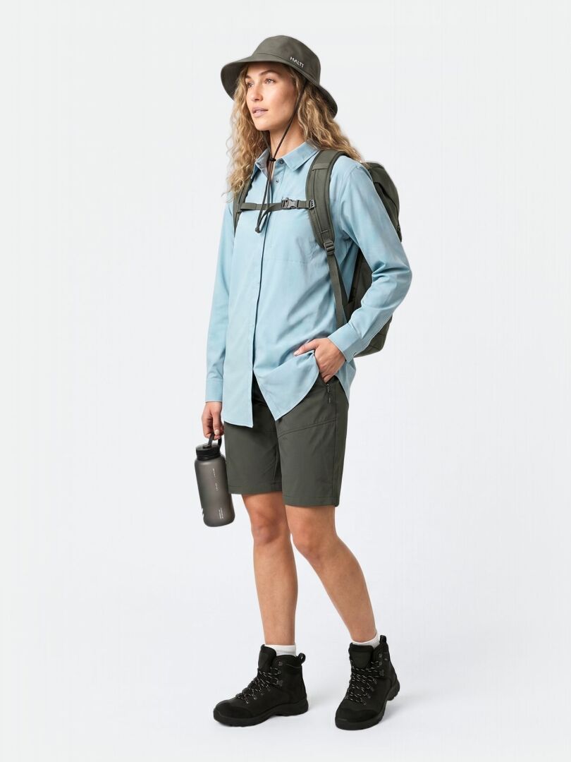 Eine Person trägt das Halti Samoy Langarm Funktionshemd Damen, grüne Shorts, Stiefel, Hut und Rucksack und hält eine Wasserflasche.