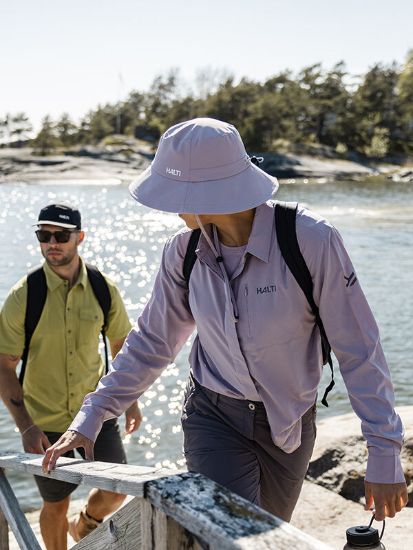 Zwei Personen, die Halti Luovi Hut UPF 90+ Eimerhüte tragen, gehen in der Sonne die Holzstufen am Wasser hinauf.