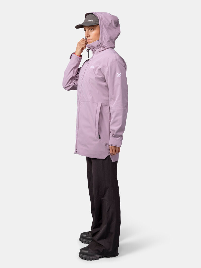 Eine Person in einem lavendelfarbenen Halti Valaja DX Parkajacke Damen und einer schwarzen Hose steht seitlich vor einem schlichten Hintergrund.