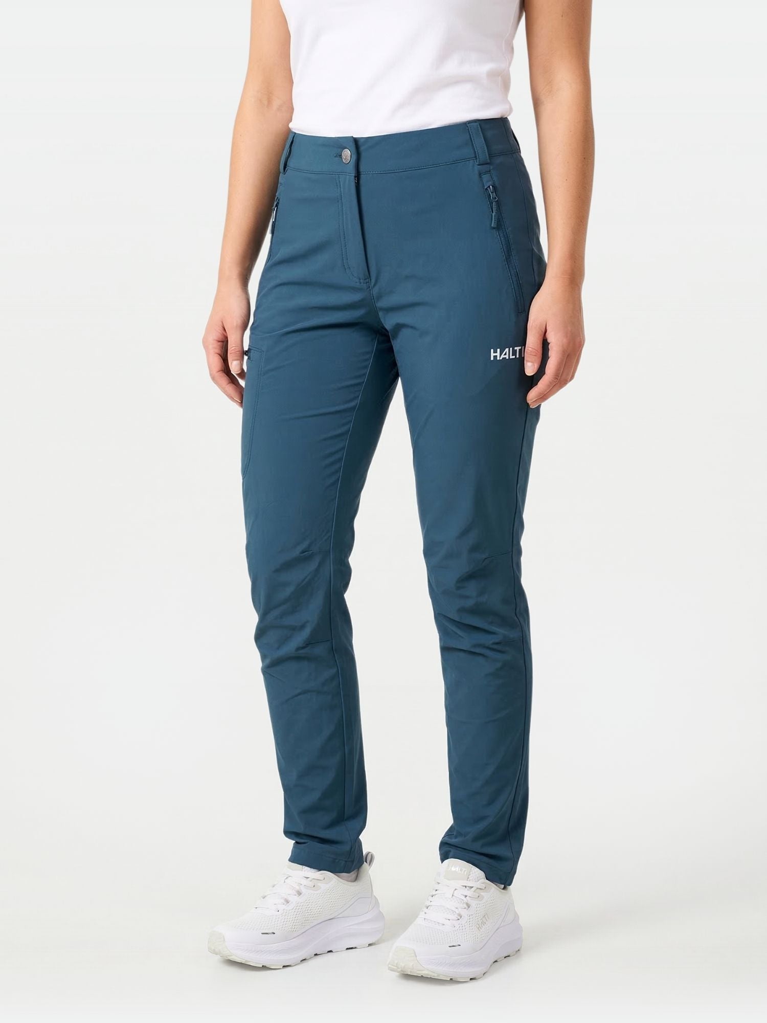 Eine Frau trägt die Halti Pallas X-stretch Hose Damen mit weißem Shirt und Sneakers vor schlichtem Hintergrund.