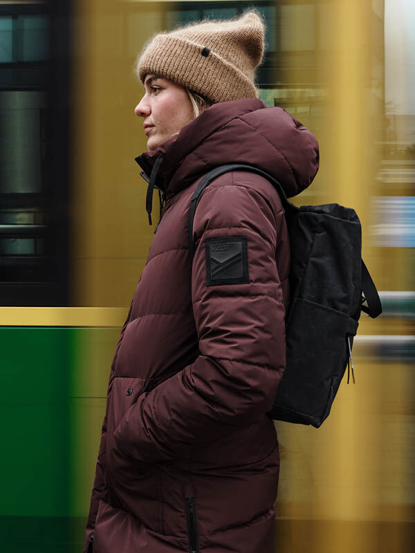 Eine Person trägt die Halti Linjat Parkajacke Damen und eine hellbraune Strickmütze vor einem verschwommenen gelben und grünen Hintergrund.