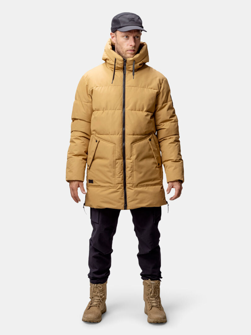 Ein Mann in einer hellbraunen Halti Linjat Parka Jacke Herren mit Thermofill-Isolierung, schwarzer Hose und Stiefeln steht vor einem schlichten Hintergrund.