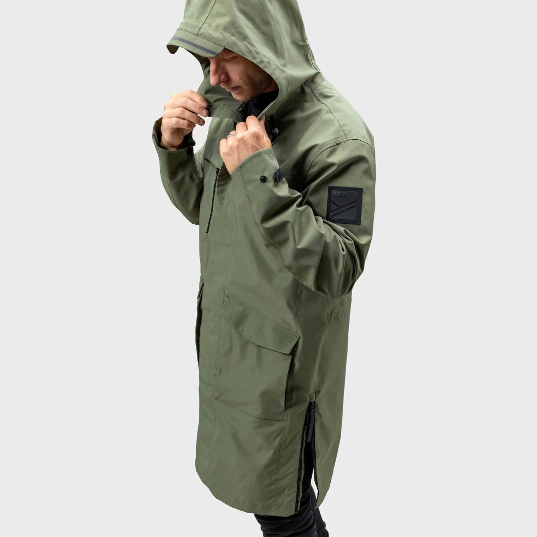 Ein Mann in einer Halti Tokoi Herren DrymaxX Parka Jacke passt die Kapuze an; wasser- und winddicht vor einem schlichten Hintergrund.