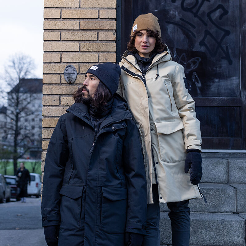Zwei Personen in Halti Bergga Damen DrymaxX Winterparkas und Beanies stehen an einer mit Graffiti bedeckten Backsteinmauer.