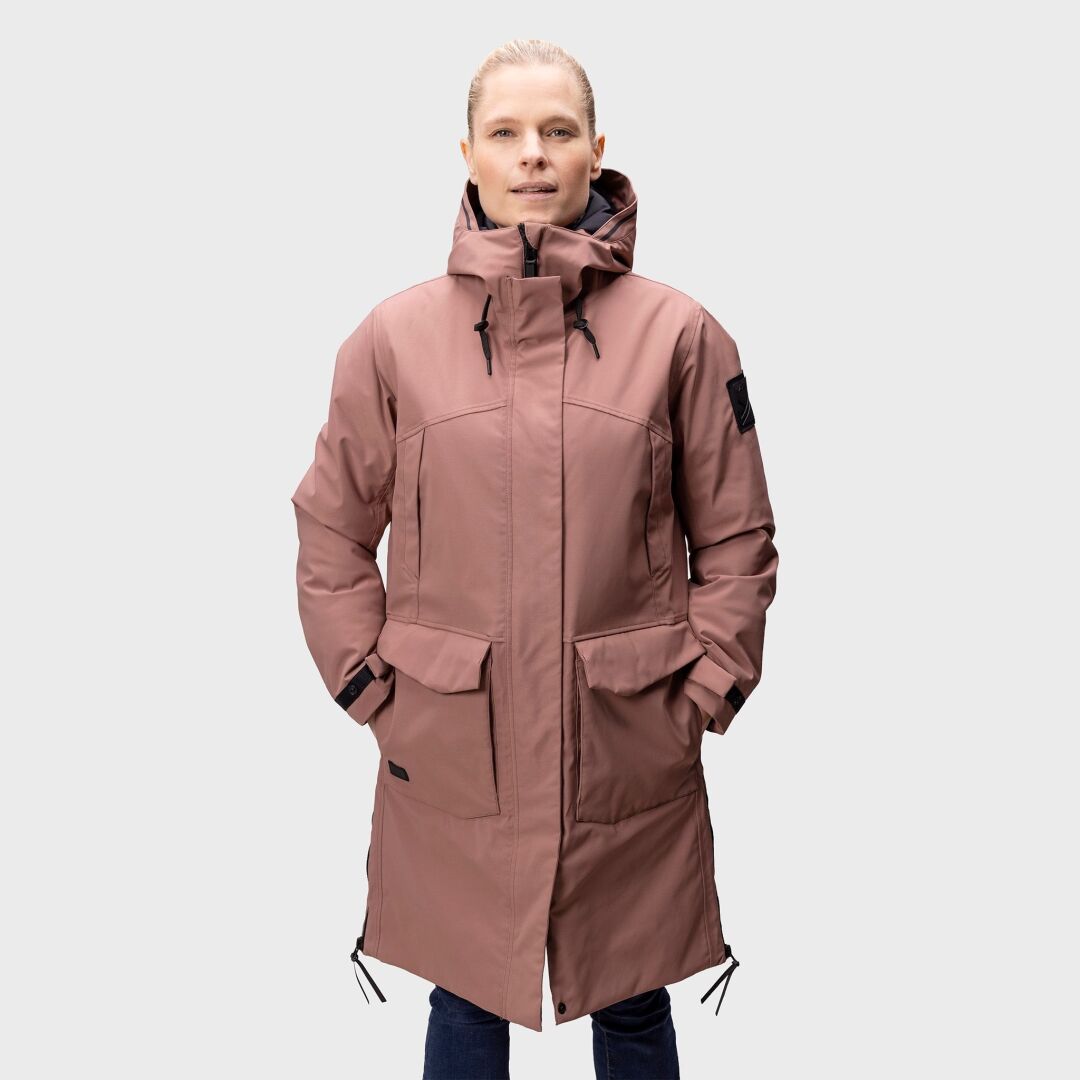 Eine Person trägt den Halti Bergga DX Winter Parka Damen in staubigem Rosa mit großen Fronttaschen vor einem schlichten Hintergrund.