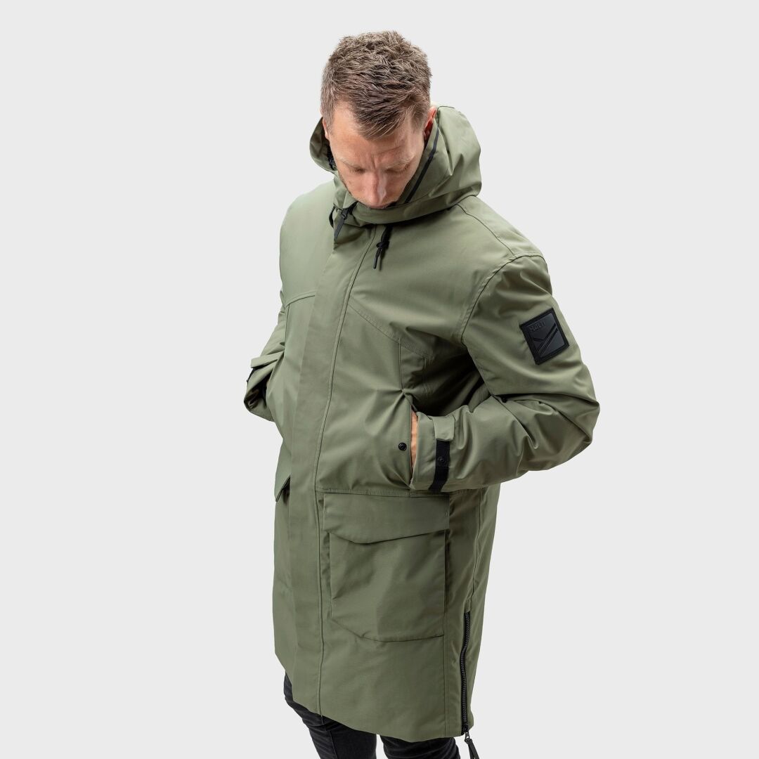 Ein Mann in einem langen, grünen Halti Bergga DX Winter Parka Herren steht mit den Händen in den Taschen vor einem schlichten, hellen Hintergrund.