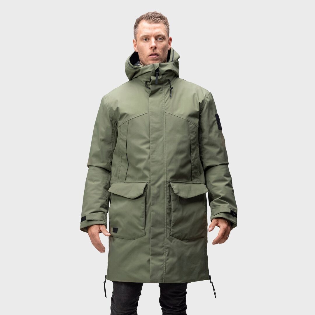 Ein Mann in einem Halti Bergga DX Winterparka mit Kapuze steht vor einem schlichten, hellgrauen Hintergrund.