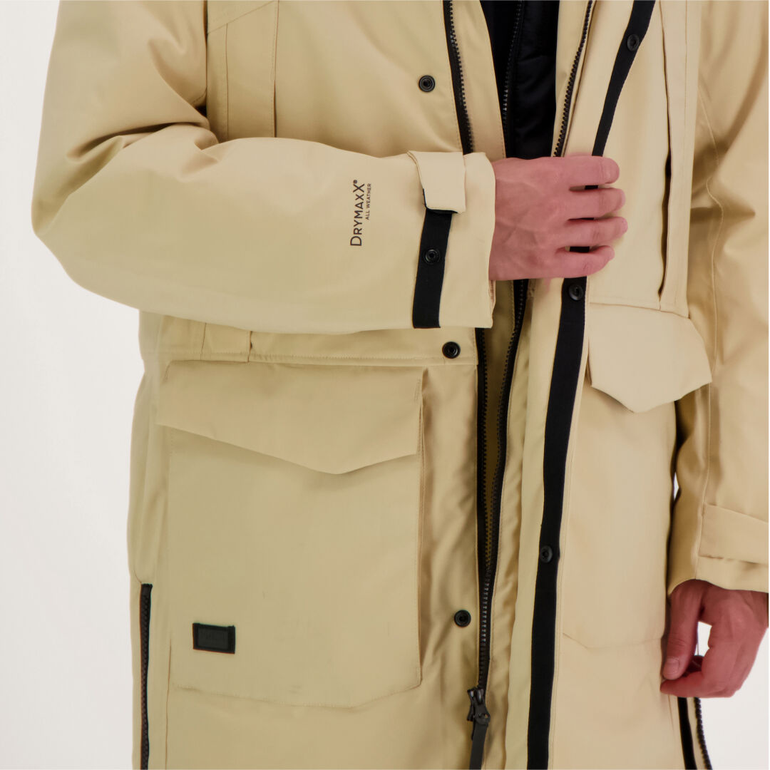 Eine Person zieht einen beigen Halti Bergga DX Winter Parka Herren mit großen Fronttaschen und DrymaxX am Ärmel auf.