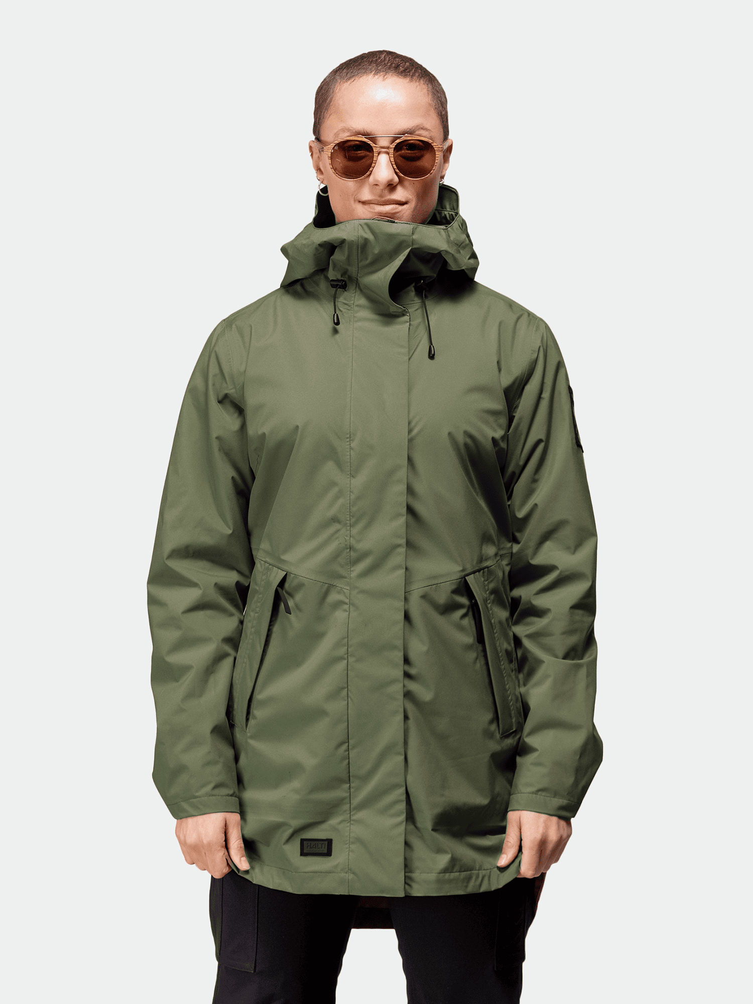 Frau trägt Halti Kurvi DrymaxX 3L Parka Jacke, dazu Sonnenbrille, vor hellem Hintergrund.