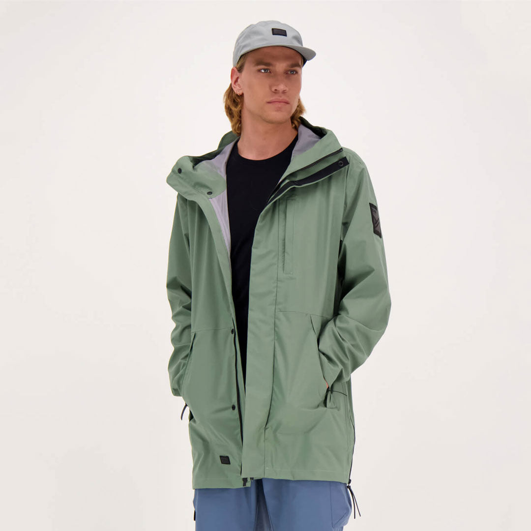 Eine Person trägt einen hellgrünen Halti Kurvi Herren DrymaxX 3L Parka, eine hellblaue Mütze, ein schwarzes Hemd und eine blaue Hose.