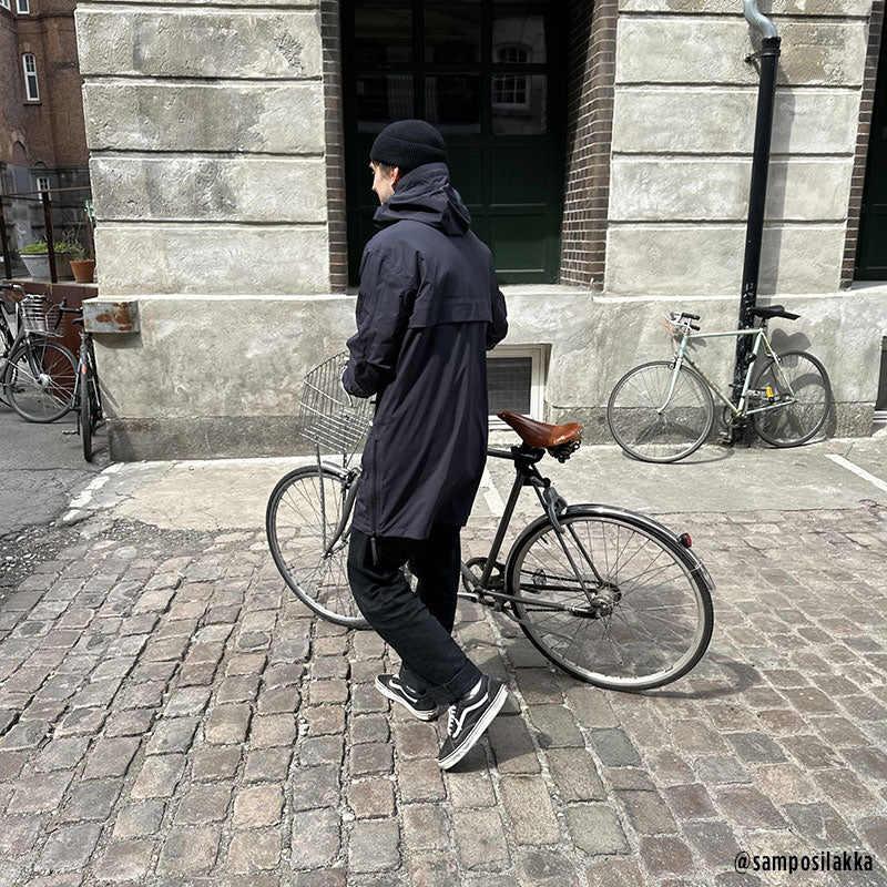 Eine Person in einer Halti Kurvi Herren DrymaxX 3L Parka Jacke schiebt ein Fahrrad auf einer Kopfsteinpflasterstraße in der Nähe geparkter Fahrräder.