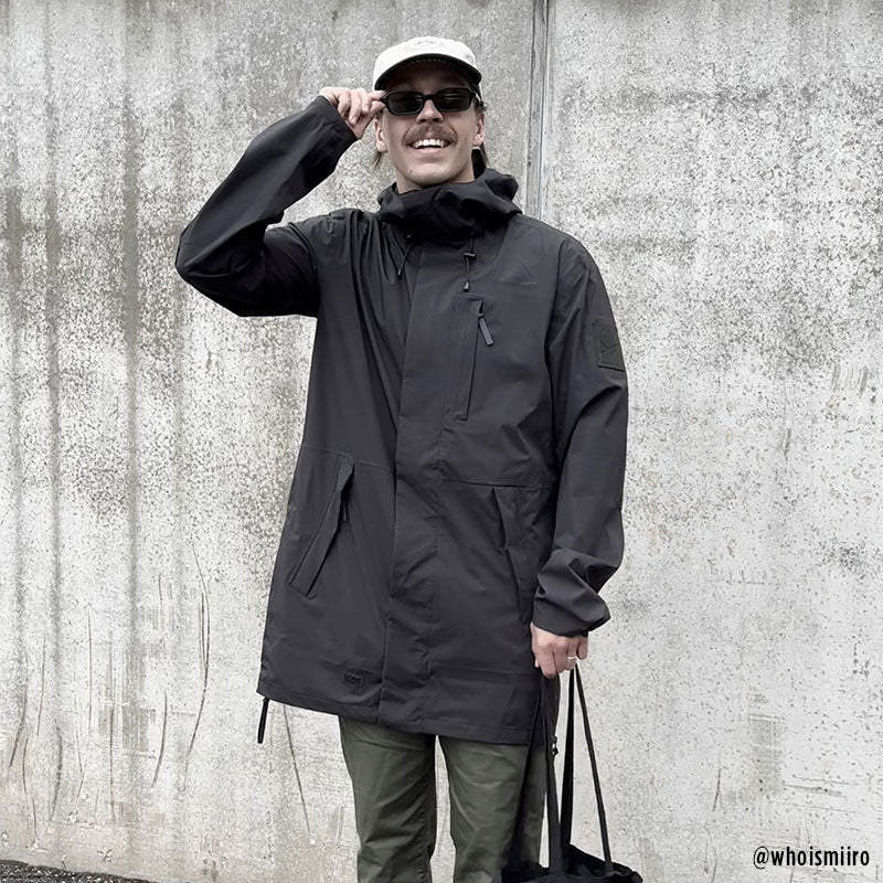 Eine Person in einem Halti Kurvi Herren DrymaxX 3L Parka, grünen Hosen, Sonnenbrille und Mütze steht mit einer Tasche an einer Betonmauer.