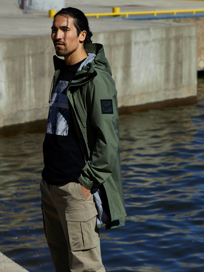 Ein Mann steht am Wasser und trägt einen grünen Halti Kurvi Herren DrymaxX 3L Parka, ein schwarzes T-Shirt mit Grafik und eine beige Cargohose.