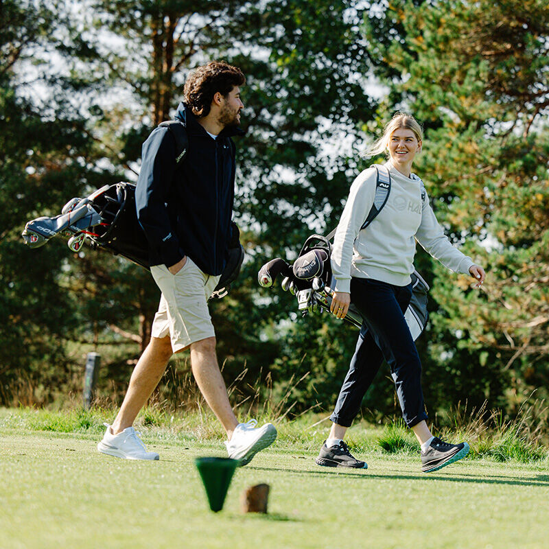 Zwei Personen auf einem Golfplatz tragen Halti Drive X-stretch Chinos Damen und tragen Golftaschen zwischen Bäumen und grünem Gras.