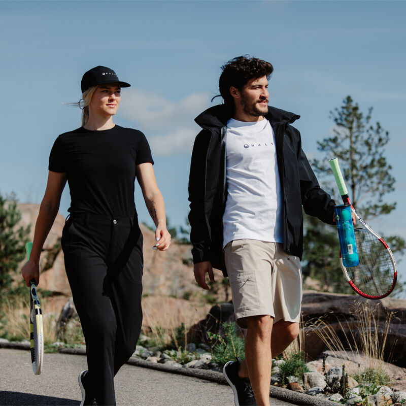 Eine Frau und ein Mann gehen mit Tennisschlägern auf einem gepflasterten Weg und tragen Halti Drive Damen X-Stretch Chinos.