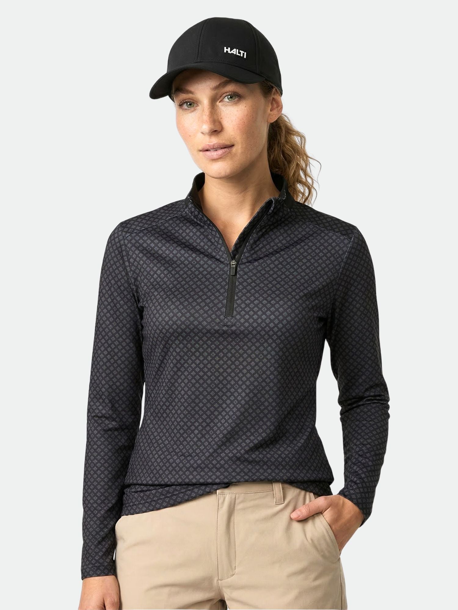 Frau mit Halti Fairway Damen Shirt mit schwarzem Muster, hellbrauner Hose und Mütze, stehend, eine Hand in der Tasche.