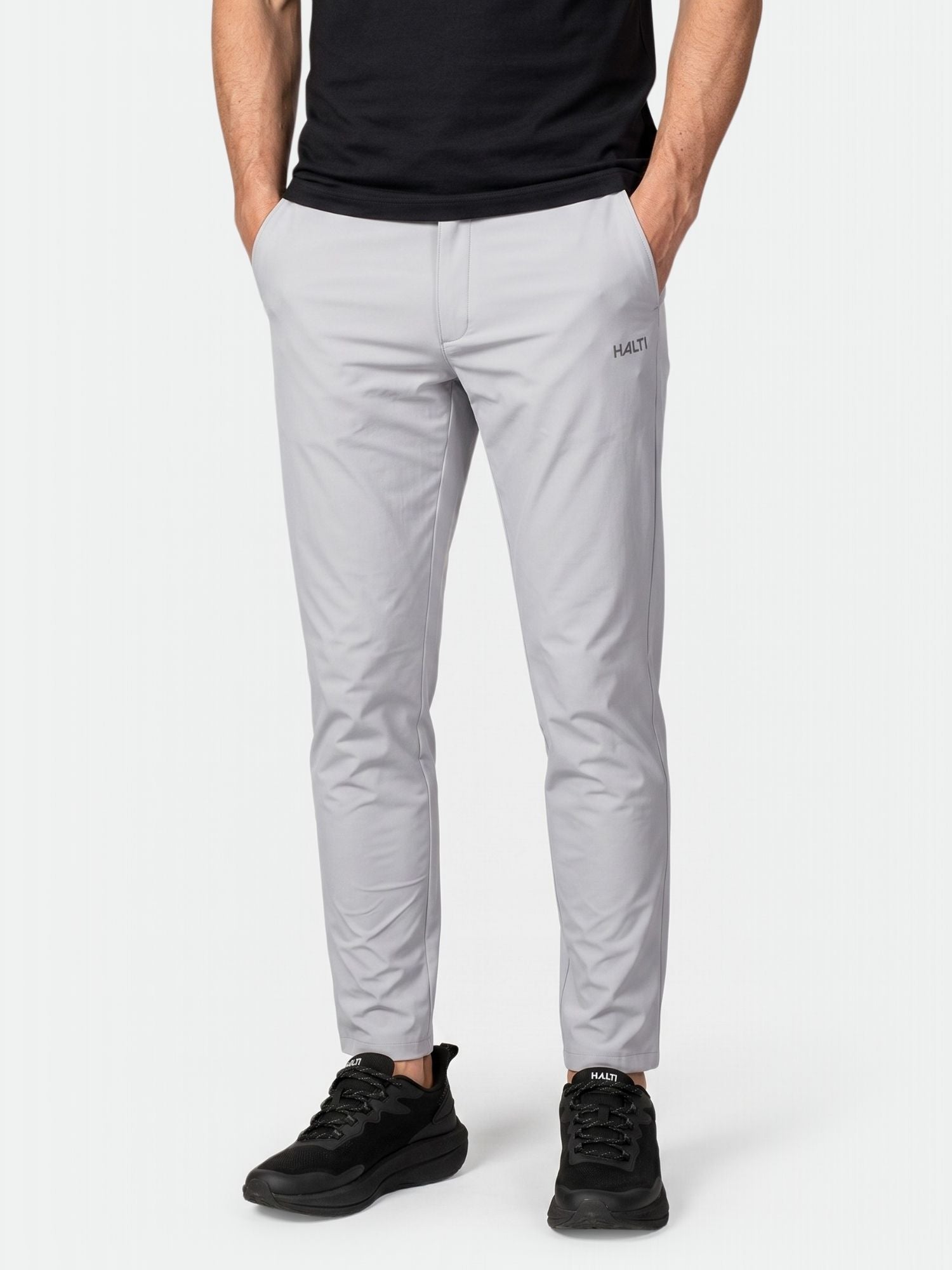 Eine Person, die eine hellgraue Halti Drive X-stretch Chinos, schwarze Turnschuhe und ein schwarzes T-Shirt trägt.