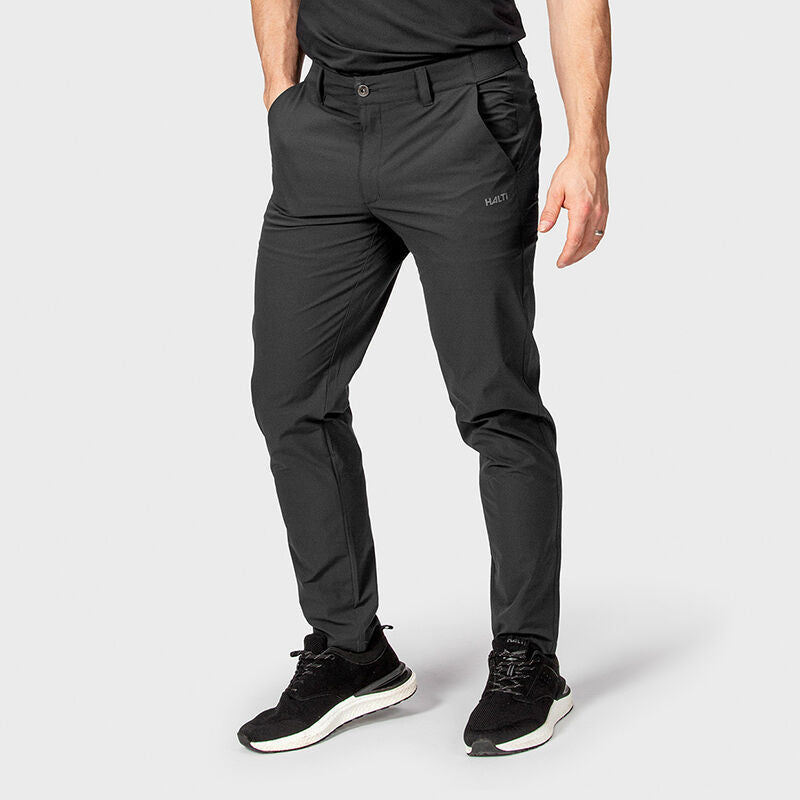 Ein Mann in schwarzen Halti Drive X-Stretch Chinos mit Turnschuhen steht mit den Händen in den Taschen vor einem schlichten Hintergrund.