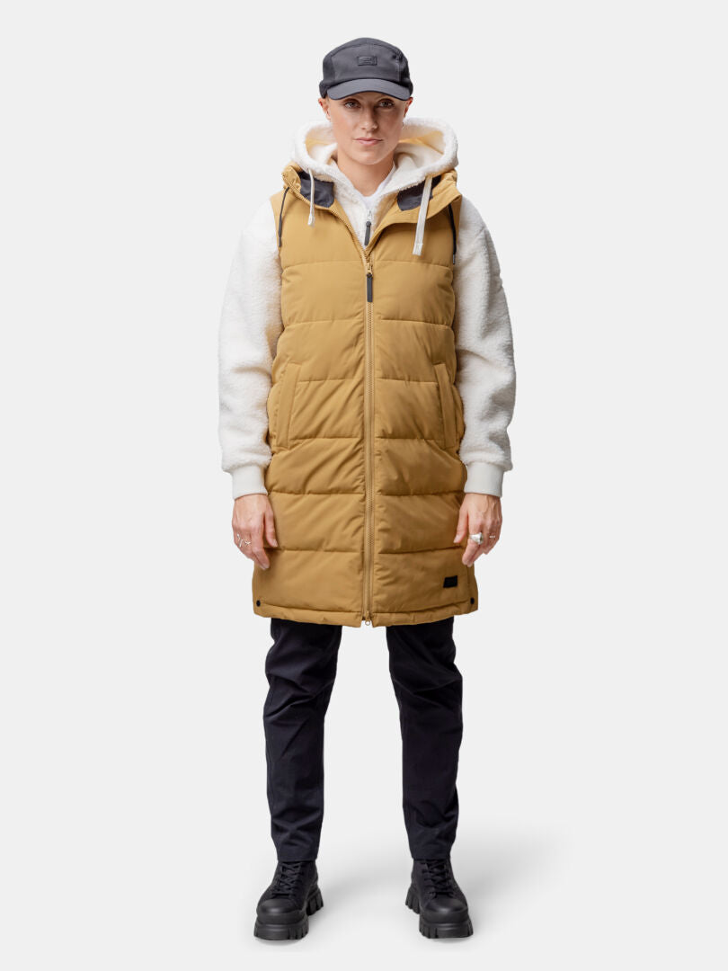 Eine Person in hellbrauner Halti Penger Puffer Weste Damen, weißem Kapuzenpullover, schwarzer Hose, Stiefeln und Mütze steht mit dem Gesicht nach vorne.