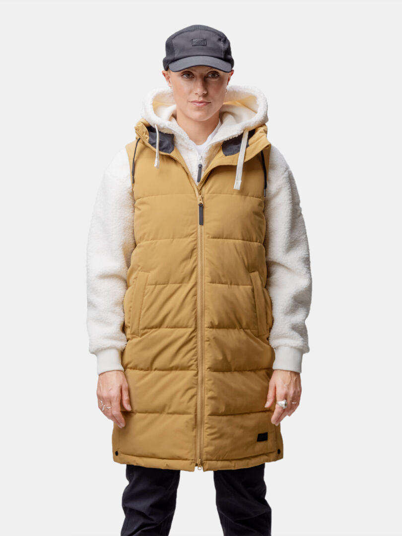 Die Person trägt eine hellbraune Halti Penger Puffer Weste Damen über einem weißen Kapuzenpulli, eine schwarze Mütze und eine Hose vor einem einfarbigen Hintergrund.