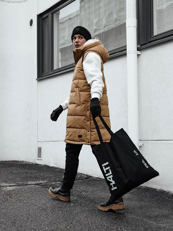 Eine Person in einer Penger Puffer Weste Damen von Halti und schwarzer Mütze geht nach draußen und trägt eine große schwarze Halti-Tasche.