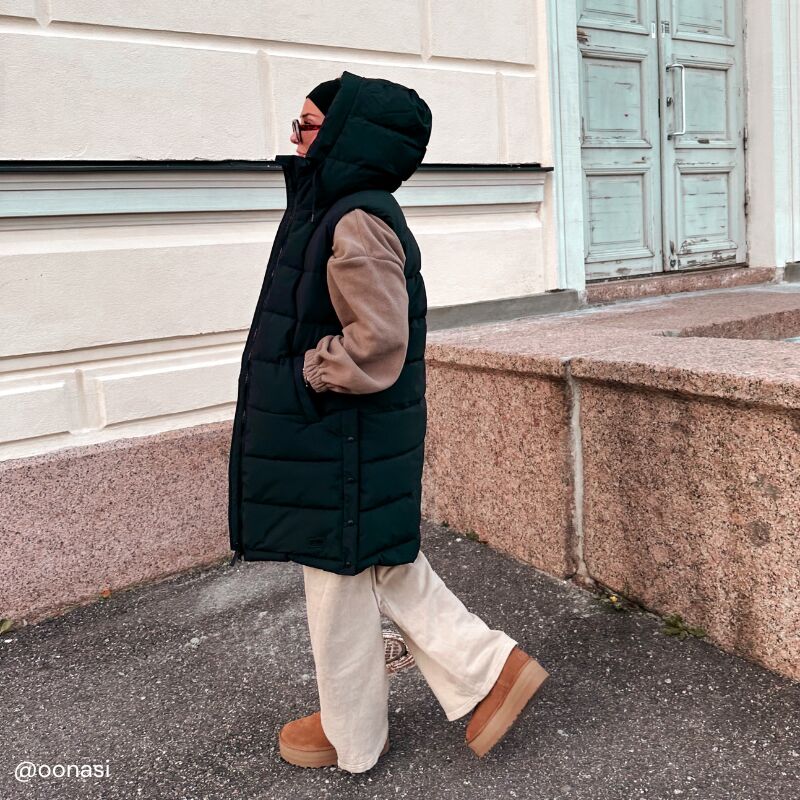 Eine Person in Halti Penger Puffer Weste Damen, beiger Hose und braunen Stiefeln geht an einer Steinmauer und einer Tür vorbei.