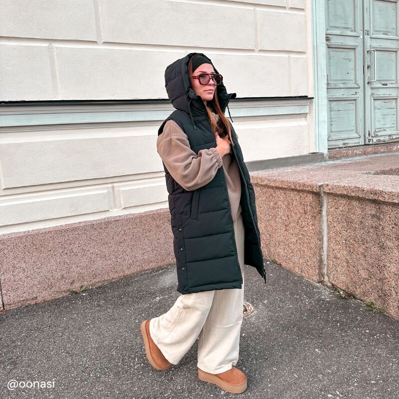 Eine Frau in Halti Penger Puffer Weste Damen, weit geschnittener Hose und Stiefeln geht an einem beigen Steingebäude vorbei.