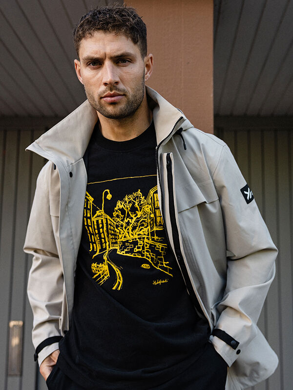 Ein Mann in einer hellen Jacke und Halti Skaltsi T-Shirt Unisex steht vor einer gestreiften Wand und schaut in die Kamera.