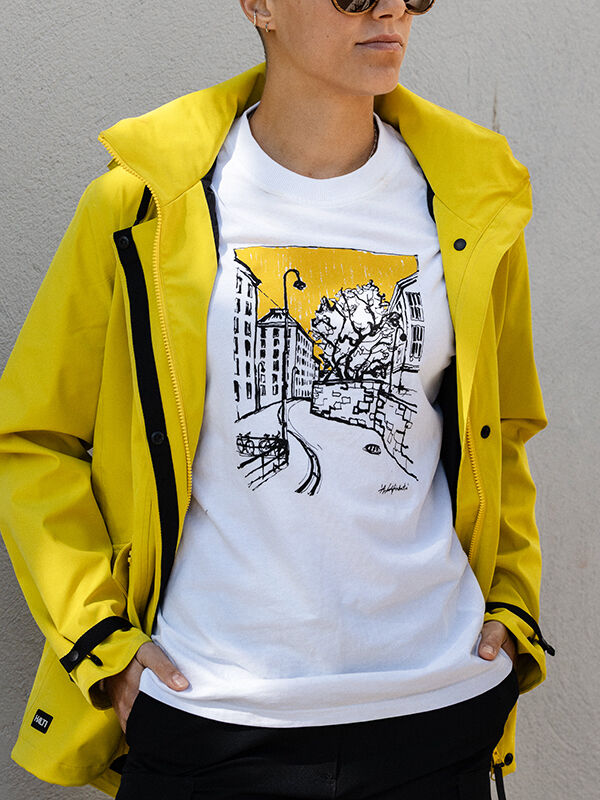 Eine Person mit Sonnenbrille, einer gelben Halti Kallio Jacke und einem Halti Skaltsi Unisex T-Shirt steht vor einer grauen Wand.