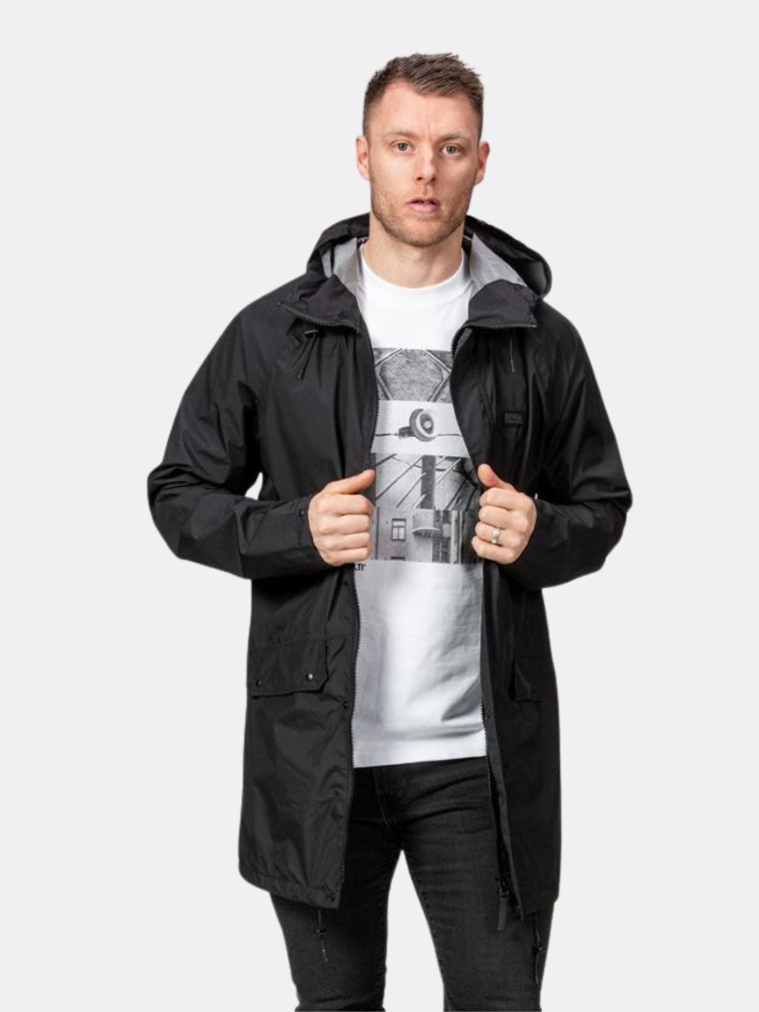 Ein Mann trägt eine schwarze Halti Kevee Parka Jacke Herren, perfekt für städtische Mobilität, mit schwarzen Jeans und einem weißen bedruckten T-Shirt.