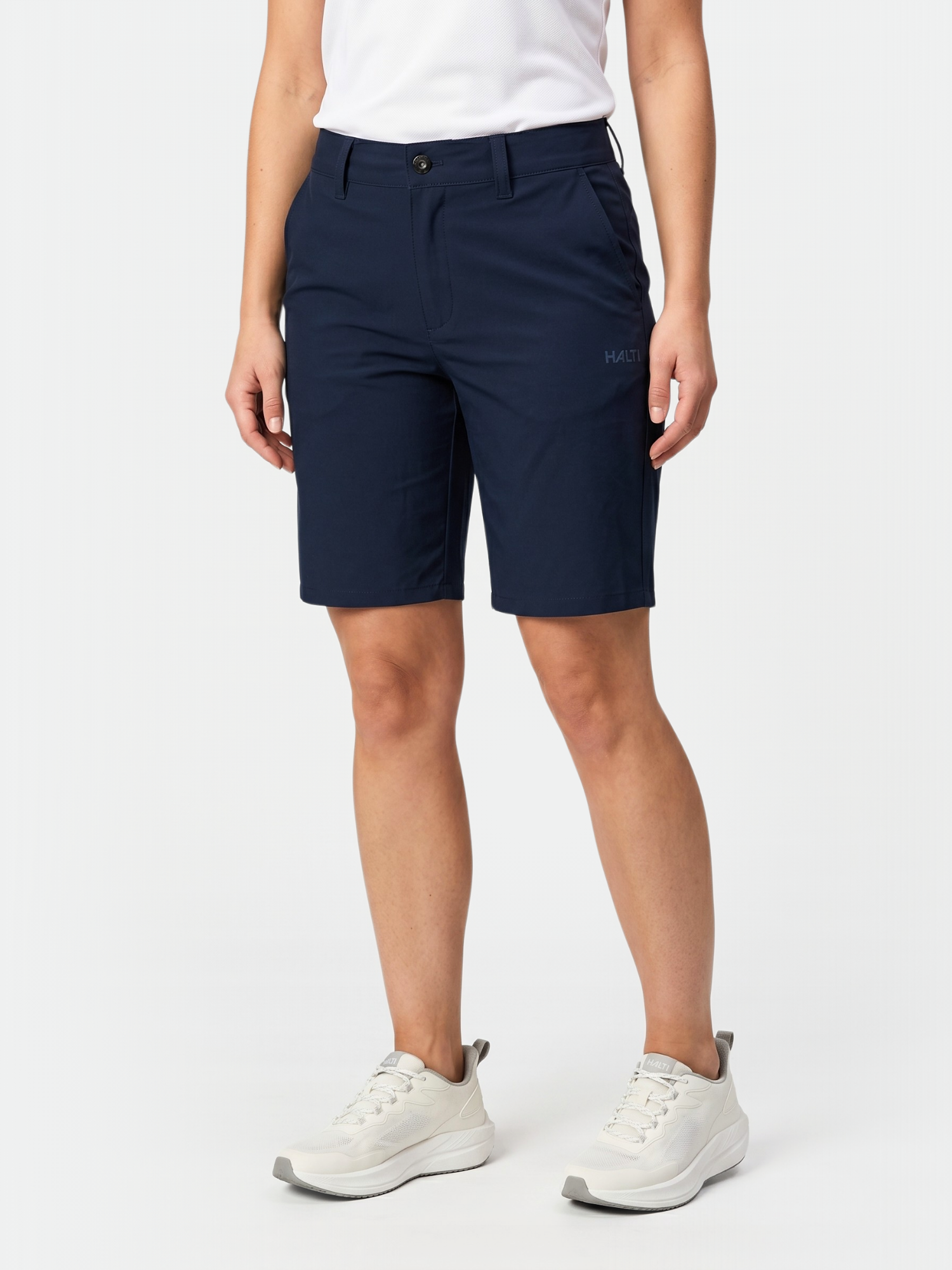 Person, die Halti Drive X-stretch Shorts Damen in navy, ein weißes Hemd und weiße Turnschuhe vor einem einfarbigen Hintergrund trägt.