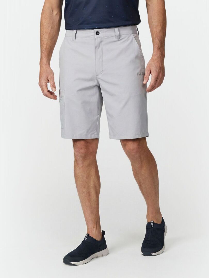 Ein Mann trägt Halti Drive Stretch Shorts Herren in Hellgrau, ein marineblaues Hemd und Slipper vor einem weißen Hintergrund.