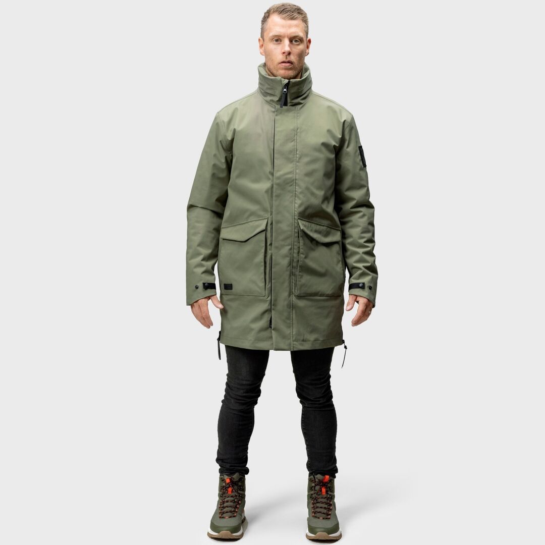 Ein Mann blickt nach vorne und trägt einen Halti Synkas 3in1 DrymaxX Parka für Herren, schwarze Hosen und graue Wanderstiefel vor einem schlichten Hintergrund.