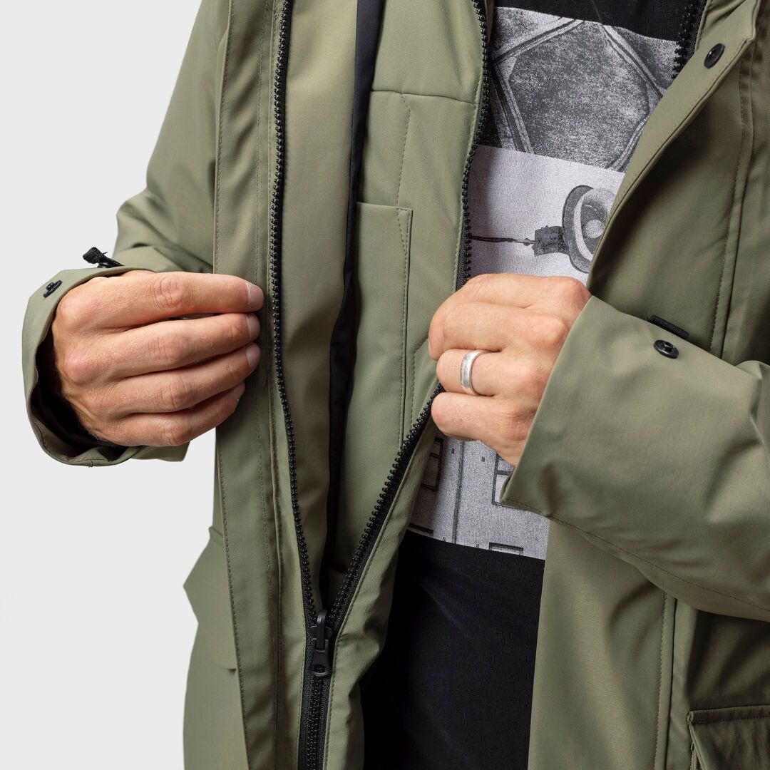 Ein Mann zieht den Reißverschluss eines olivgrünen Halti Synkas 3in1 DrymaxX Parka für Herren über einem schwarzen T-Shirt mit Kameragrafik zu.