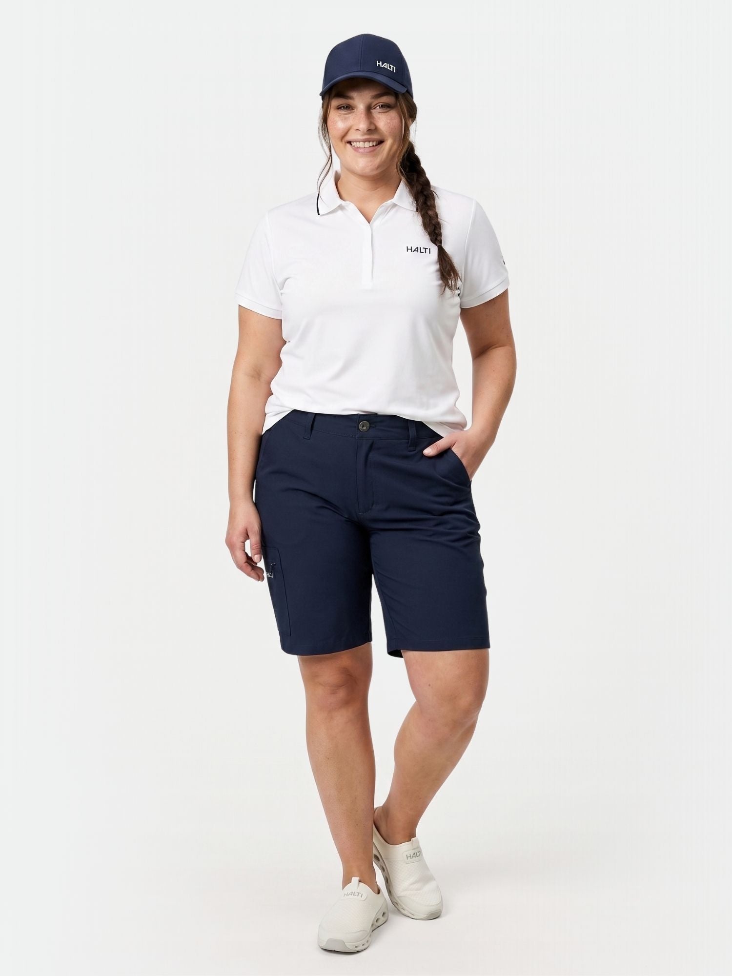 Eine Person mit marineblauer Kappe, weißem Polo und Halti Drive X-stretch Shorts Damen Plus steht vor einem schlichten weißen Hintergrund.