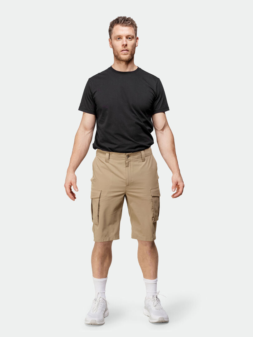 Der Mann steht mit Blick nach vorne da und trägt ein schwarzes T-Shirt, khakifarbene Cargo-Shorts von Halti Roams Shorts für Herren, weiße Socken und Turnschuhe.