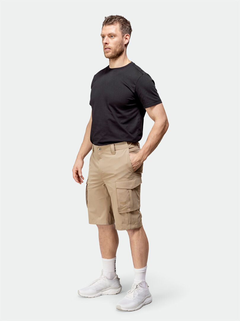 Ein Mann in einem schwarzen T-Shirt und beigen Halti Roams Shorts Herren posiert vor einem schlichten Hintergrund, mit weißen Socken und Turnschuhen.