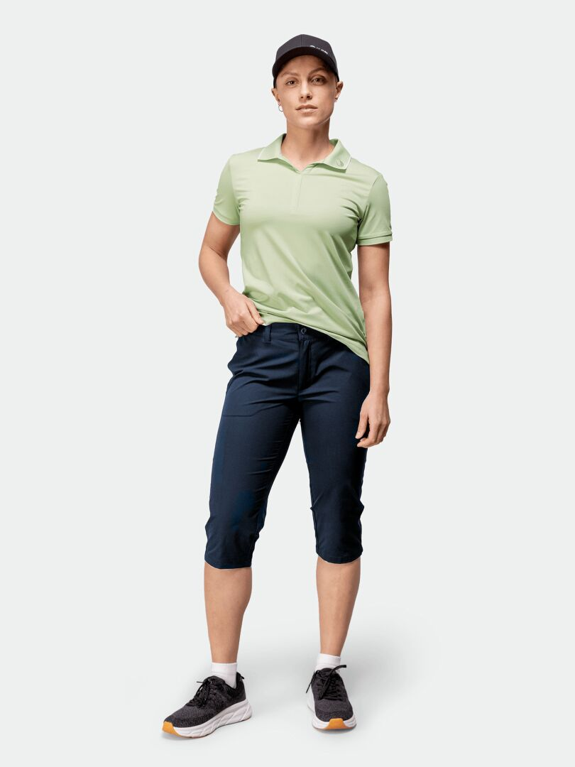 Eine Person trägt Halti Drive X-stretch Caprihose Damen, ein hellgrünes Polo, eine schwarze Mütze und schwarze Turnschuhe mit weißen Sohlen.