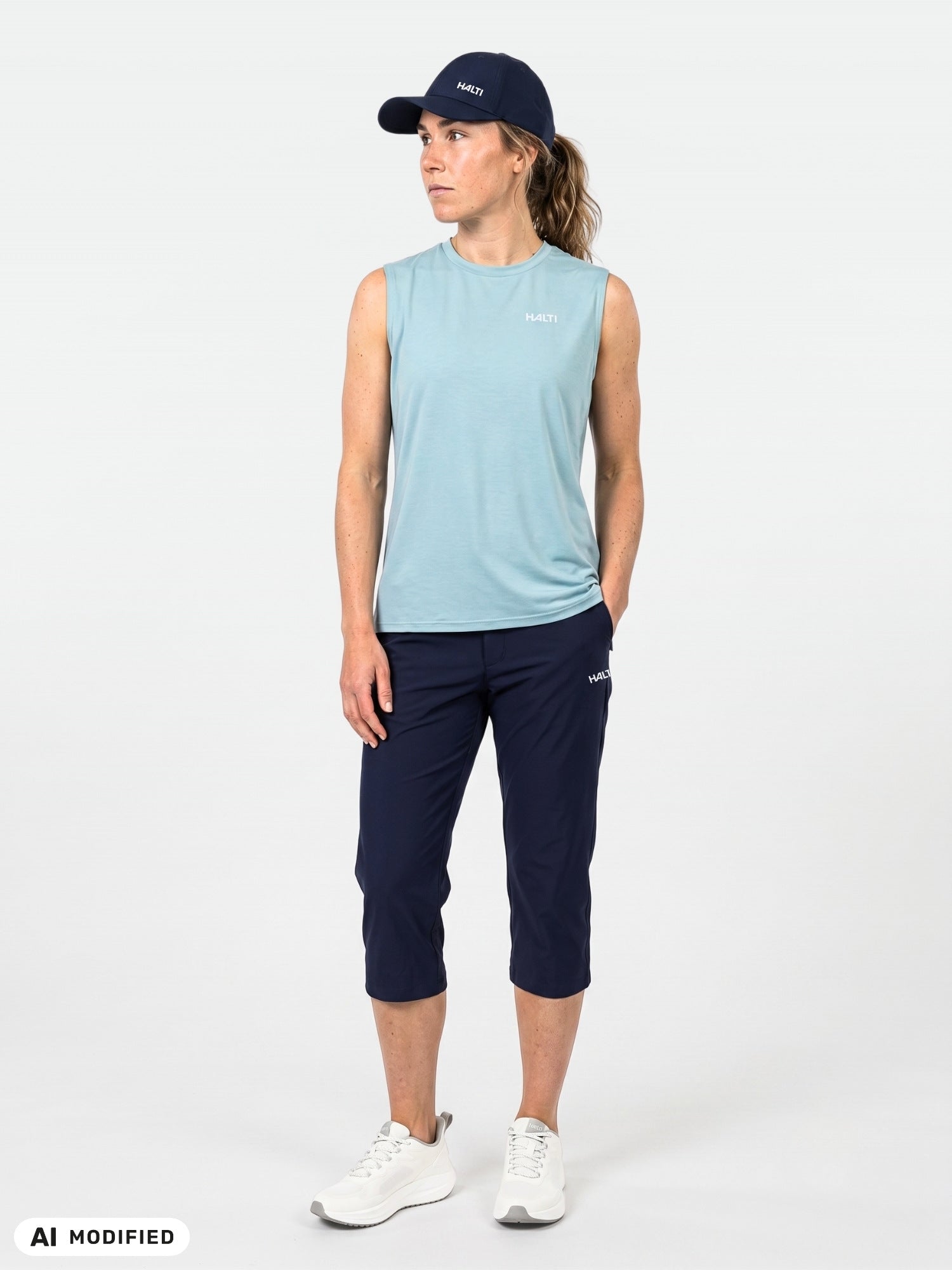 Frau in einer Caprihose „Halti Drive X-Stretch“ für Damen, blaues, ärmelloses Oberteil, marineblaue Mütze und weiße Sneaker vor einfarbigem Hintergrund.
