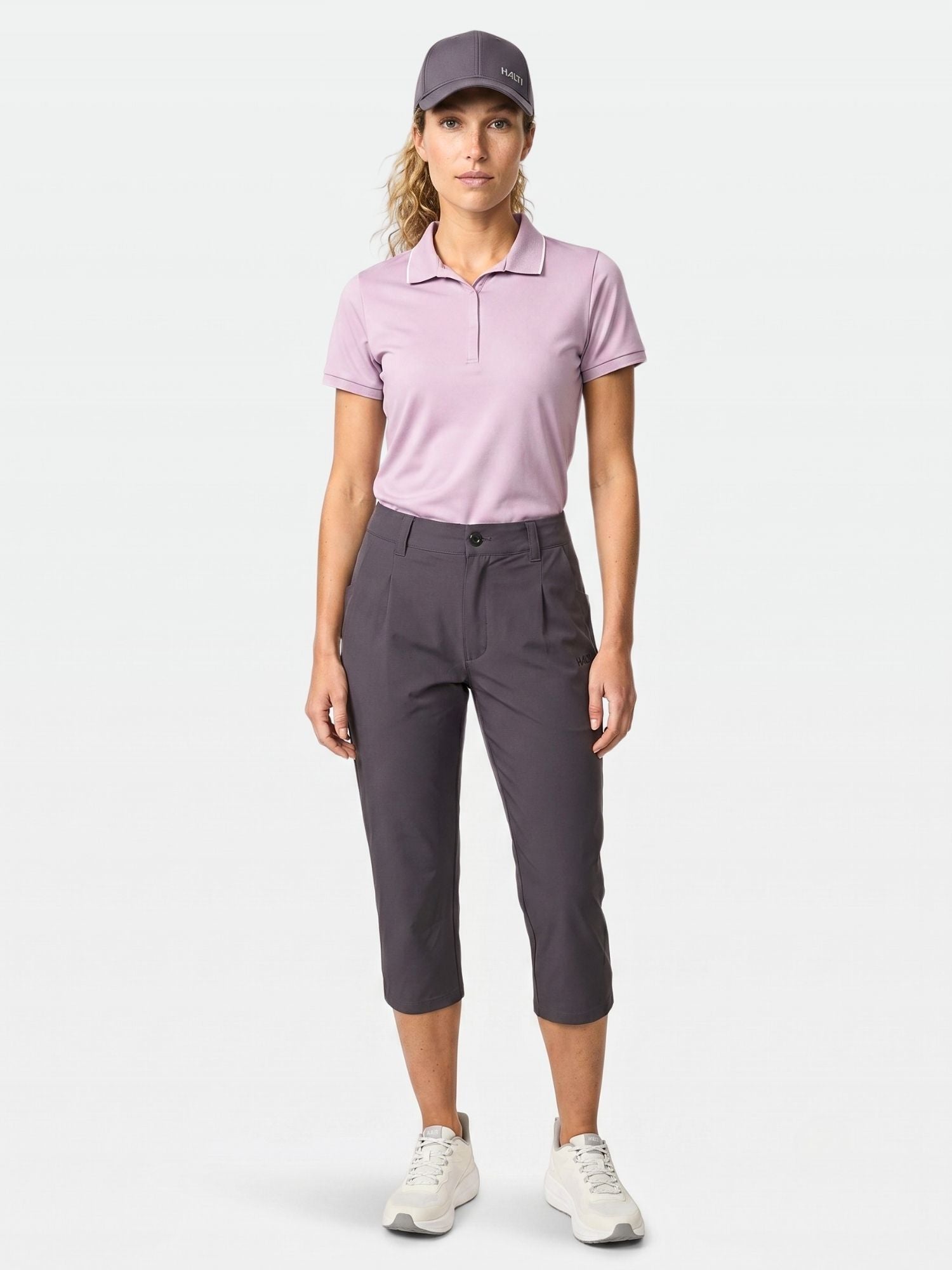 Eine Person sieht nach vorne und trägt Halti Drive X-stretch Caprihose Damen, eine graue Mütze, ein rosa Polo und weiße Turnschuhe.