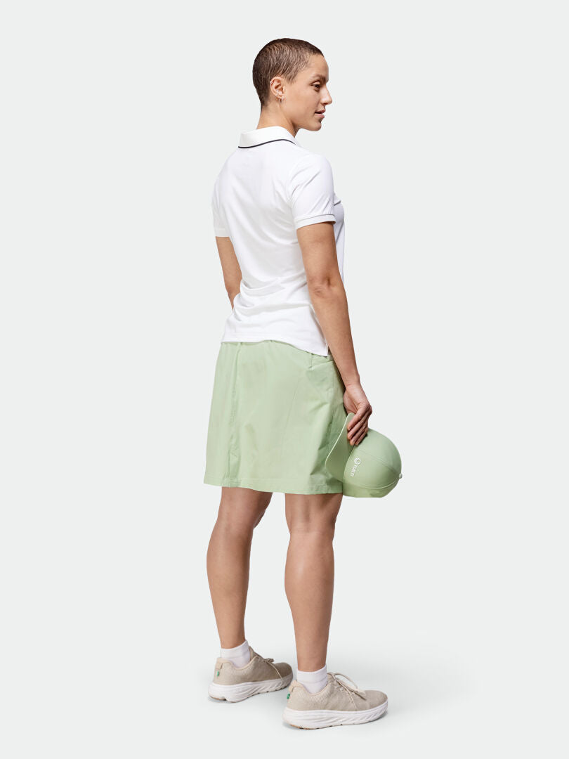 Abgewandte Frau in weißem Poloshirt, hellgrünem Halti Drive X-Stretch-Skort und Turnschuhen, bereit für Outdoor-Aktivitäten.