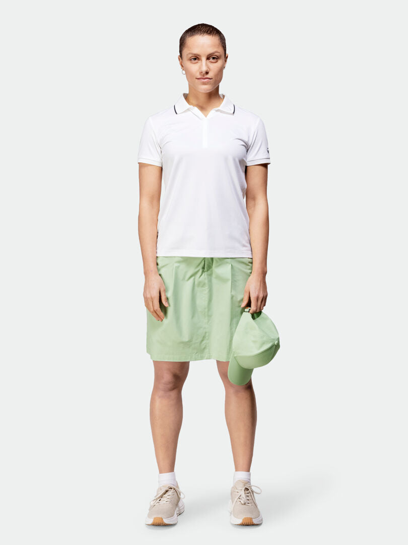 Eine Person steht mit Blick nach vorne, trägt ein weißes Poloshirt, einen Halti Drive X-Stretch Skort Damen, Turnschuhe und hält eine grüne Kappe.