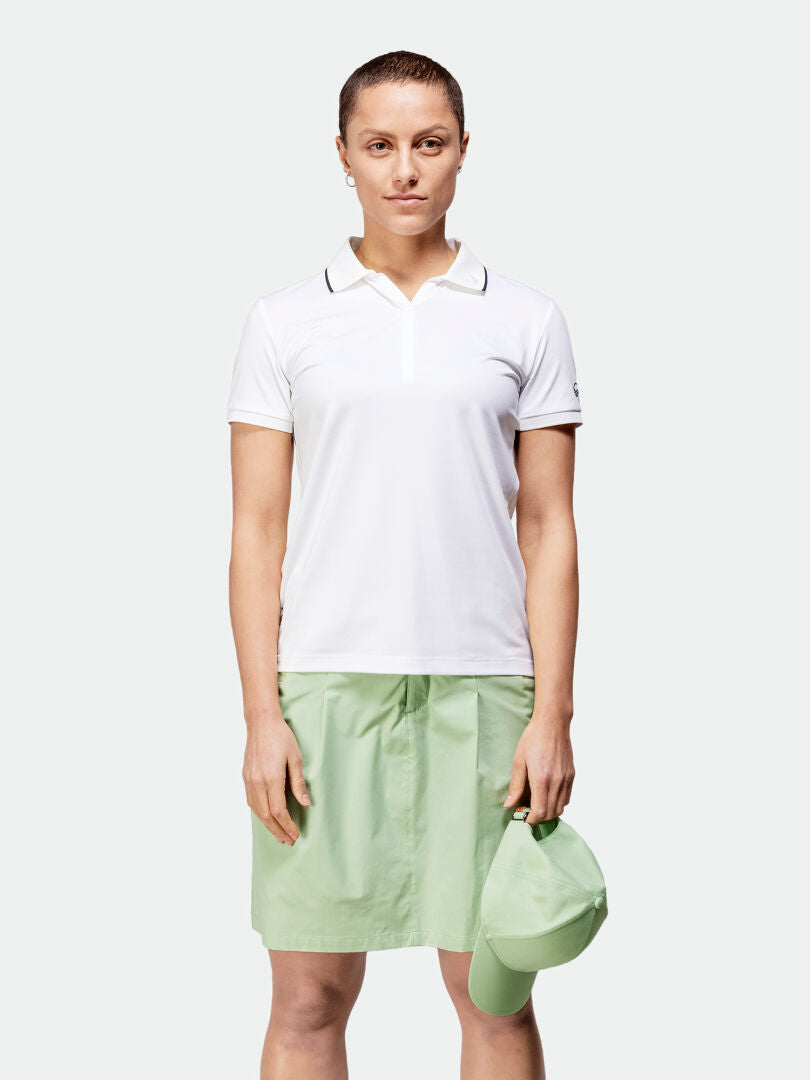 Eine Frau in einem weißen Poloshirt und einem Halti Drive X-Stretch-Skort hält eine passende Mütze und ist bereit für Outdoor-Aktivitäten.