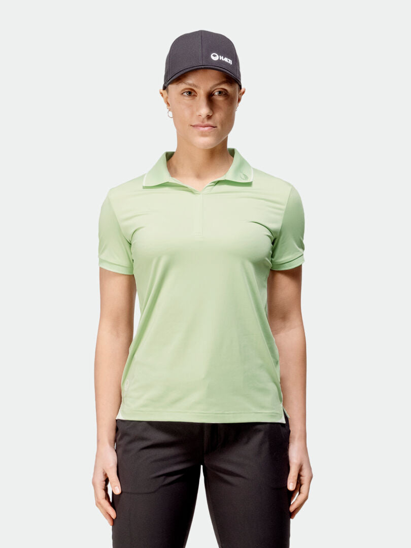 Eine Person in einem hellgrünen Halti Swingi Technishes Poloshirt für Damen, schwarzen Hosen und einer grauen Mütze steht vor einem schlichten Hintergrund.