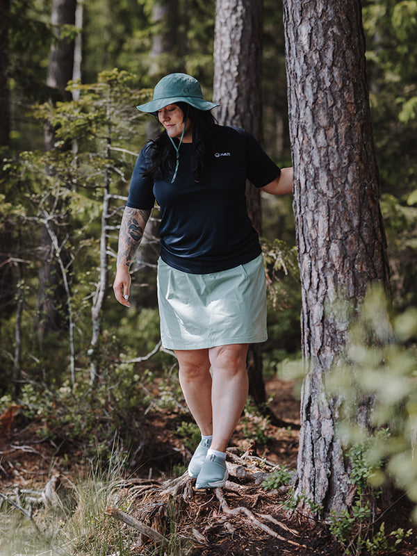 Eine Person in Outdoor-Kleidung und Turnschuhen trägt Halti Drive X-Stretch Rock Damen Plus Shorts, einen Hut und ein schwarzes Shirt neben einem Baum.