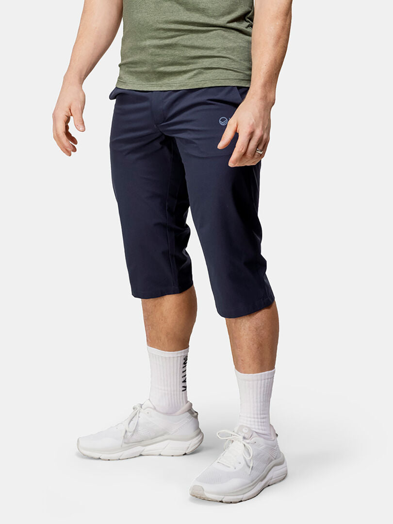 Eine Person in Halti Drive X-stretch Caprihose Herren, weißen Ringelsocken und Turnschuhen steht vor einem schlichten Hintergrund.