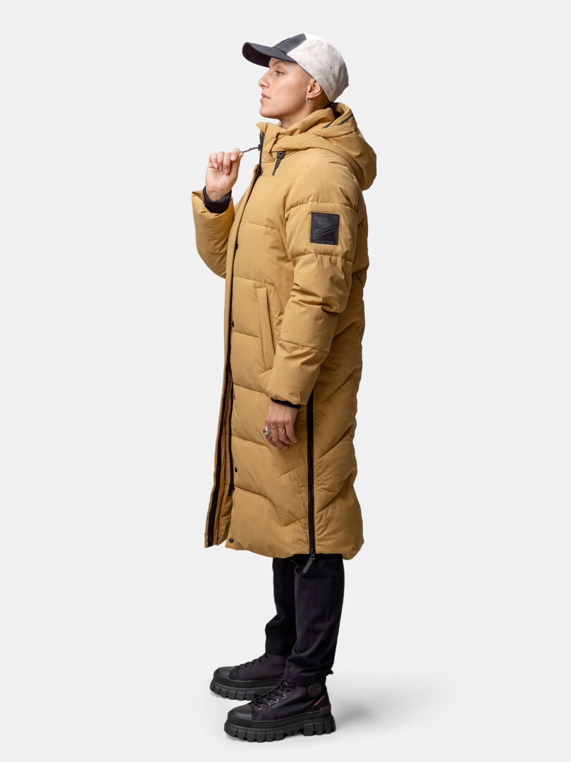 Eine Frau in einer hellbraunen Halti Brahe Puffer Long Winterjacke, einer schwarzen Hose und einer Mütze steht nach links.