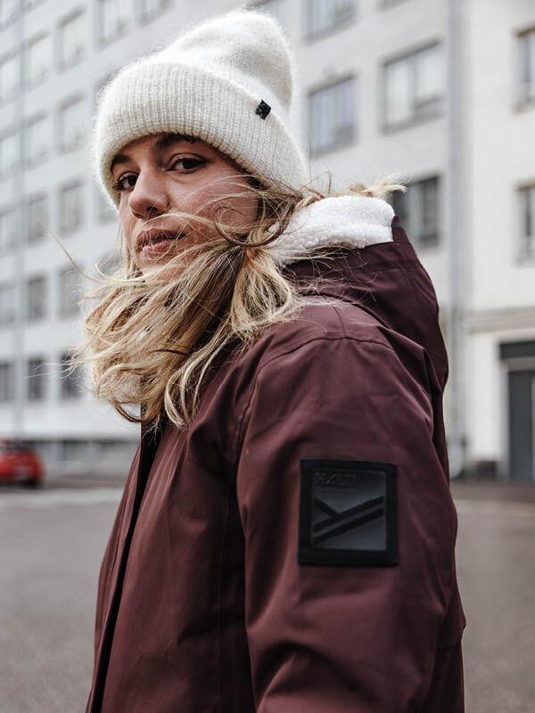 Eine Person in kastanienbrauner Jacke und Halti Vuolle Mütze steht auf einer Stadtstraße und blickt in die Kamera.